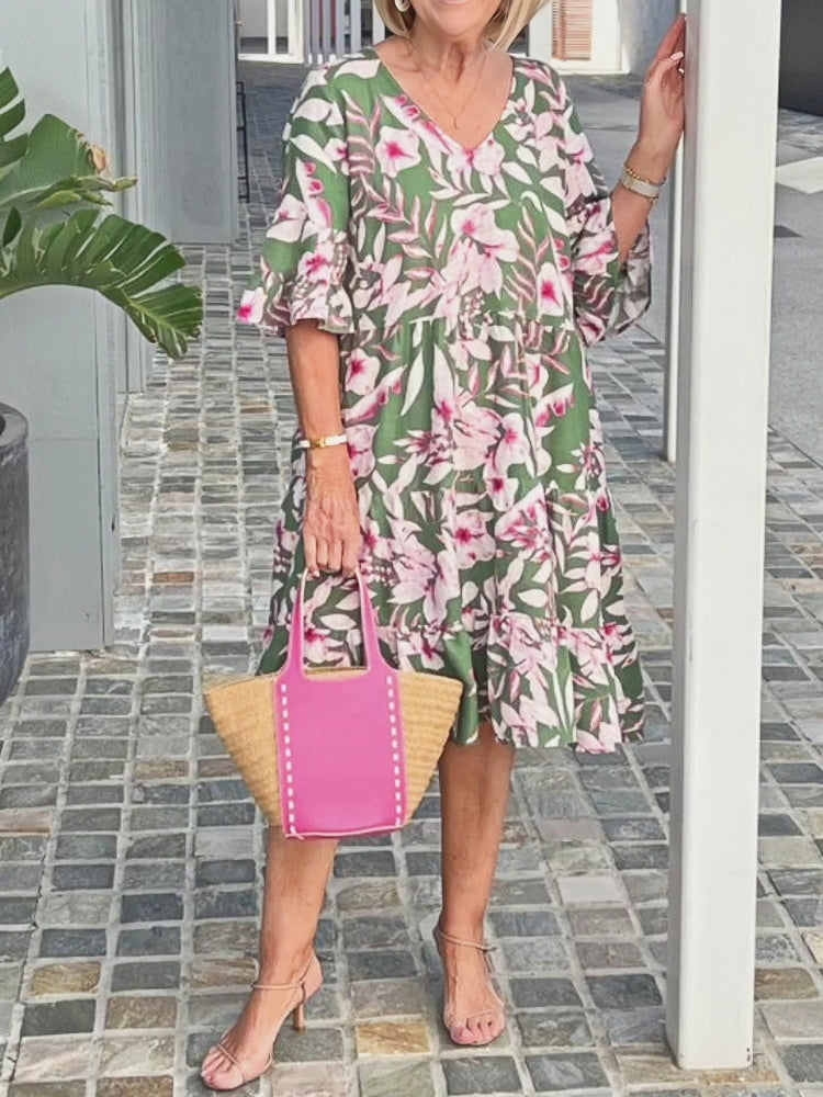 A woman wearing a blue and white speckled print midi dress with a V-neckline, elbow-length flutter sleeves, and a tiered skirt, holding a woven straw tote bag.