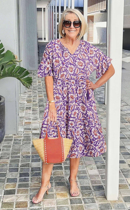 A woman wearing a blue and white speckled print midi dress with a V-neckline, elbow-length flutter sleeves, and a tiered skirt, holding a woven straw tote bag.