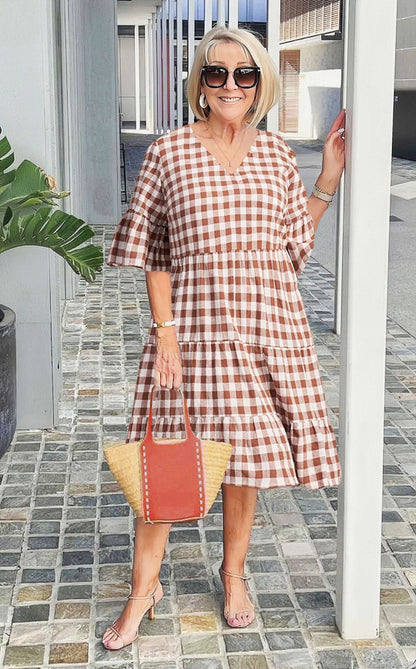 A woman wearing a blue and white speckled print midi dress with a V-neckline, elbow-length flutter sleeves, and a tiered skirt, holding a woven straw tote bag.