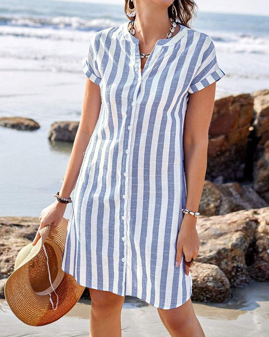 Blue / White - A woman wearing a blue and white vertically striped mini shirt dress with cuffed short sleeves and a button-down front, standing on a beach holding a straw hat.