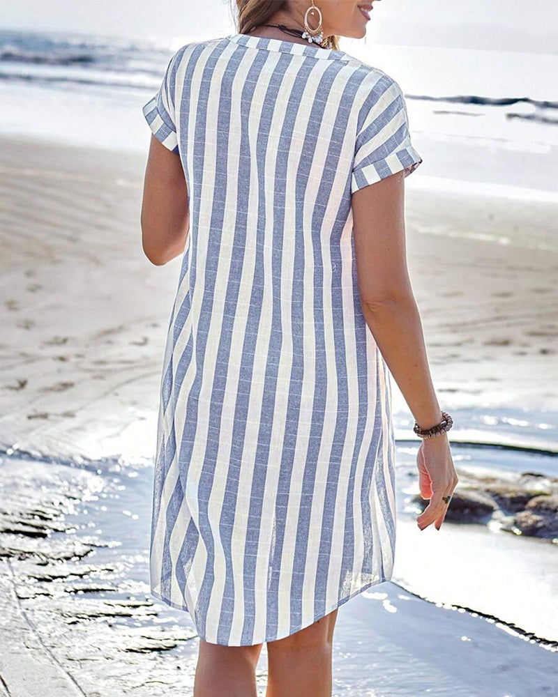 Blue / White - A woman wearing a blue and white vertically striped short-sleeve shirt dress with a split neckline, standing on a beach holding a straw sunhat.