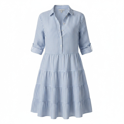 Green - A woman wearing a yellow and white vertical striped shirt dress with a collared neckline, button-down front, three-quarter sleeves, and a tiered skirt, standing on a sunlit cobblestone street.