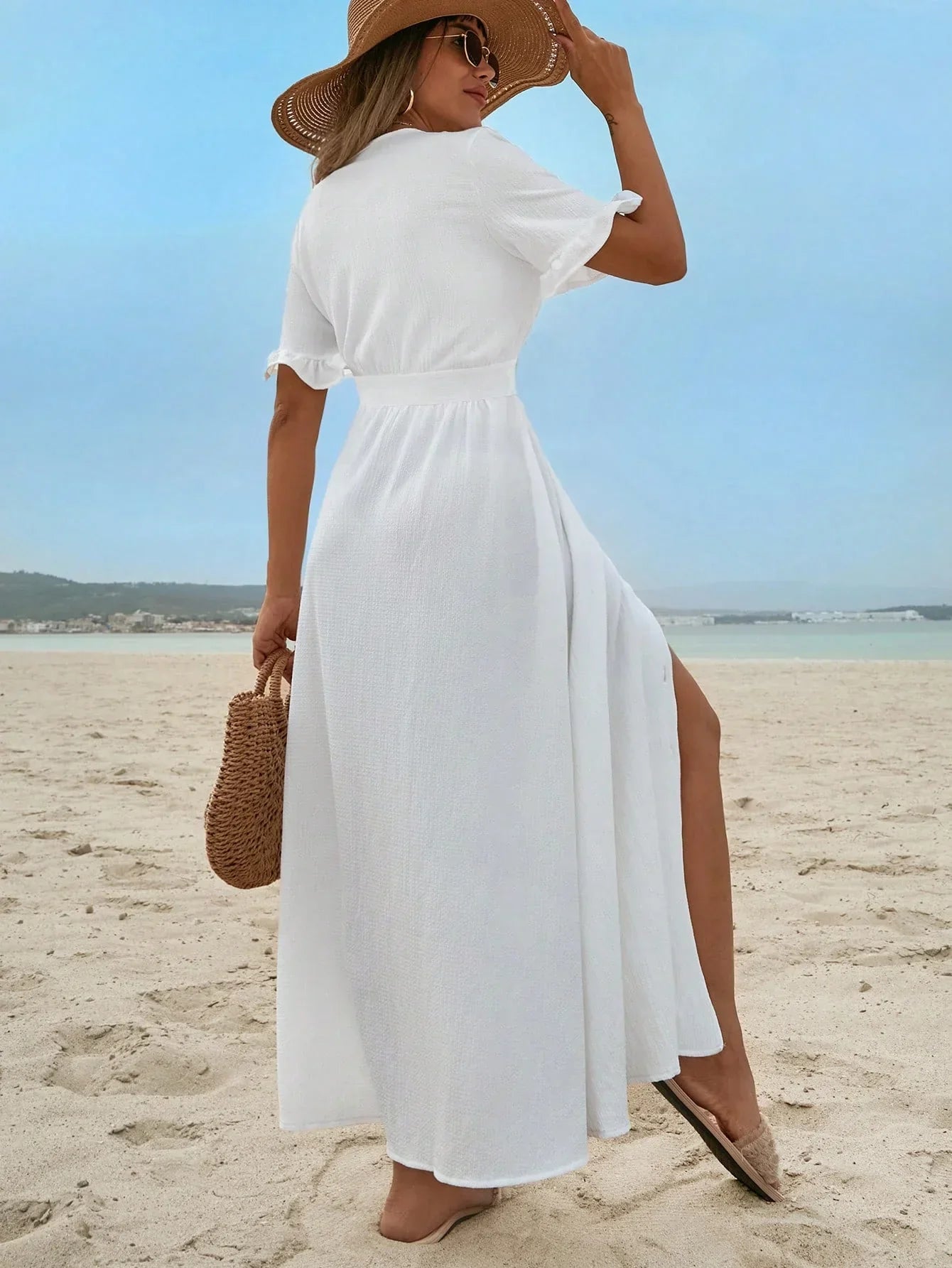 White - A woman on a sandy beach wearing a white textured crepe maxi dress with a V-neckline, flutter sleeves, button-down front, and a high leg slit, holding a woven straw handbag.