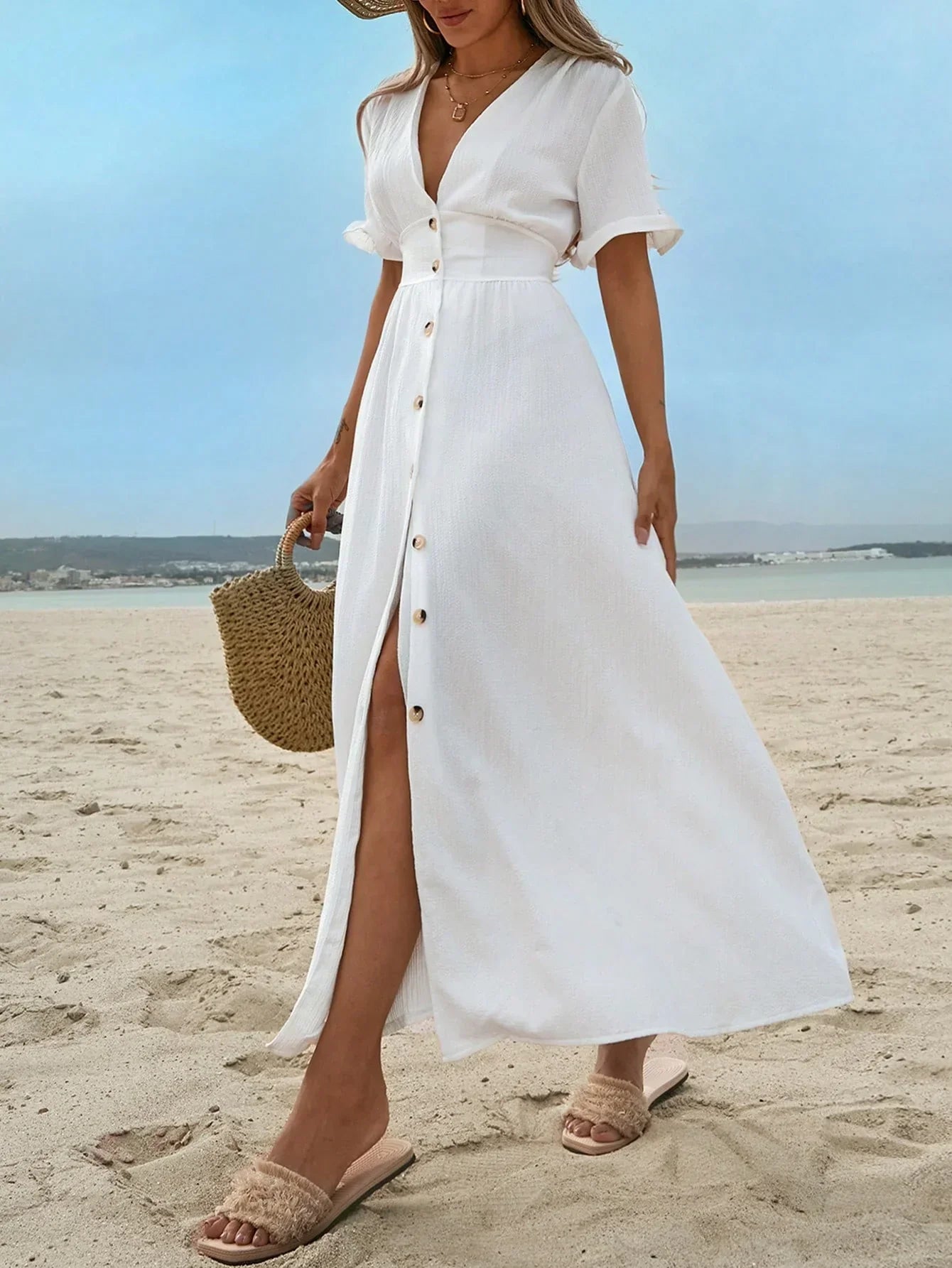 White - A woman on a sandy beach wearing a white textured crepe maxi dress with a V-neckline, flutter sleeves, button-down front, and a high leg slit, holding a woven straw handbag.