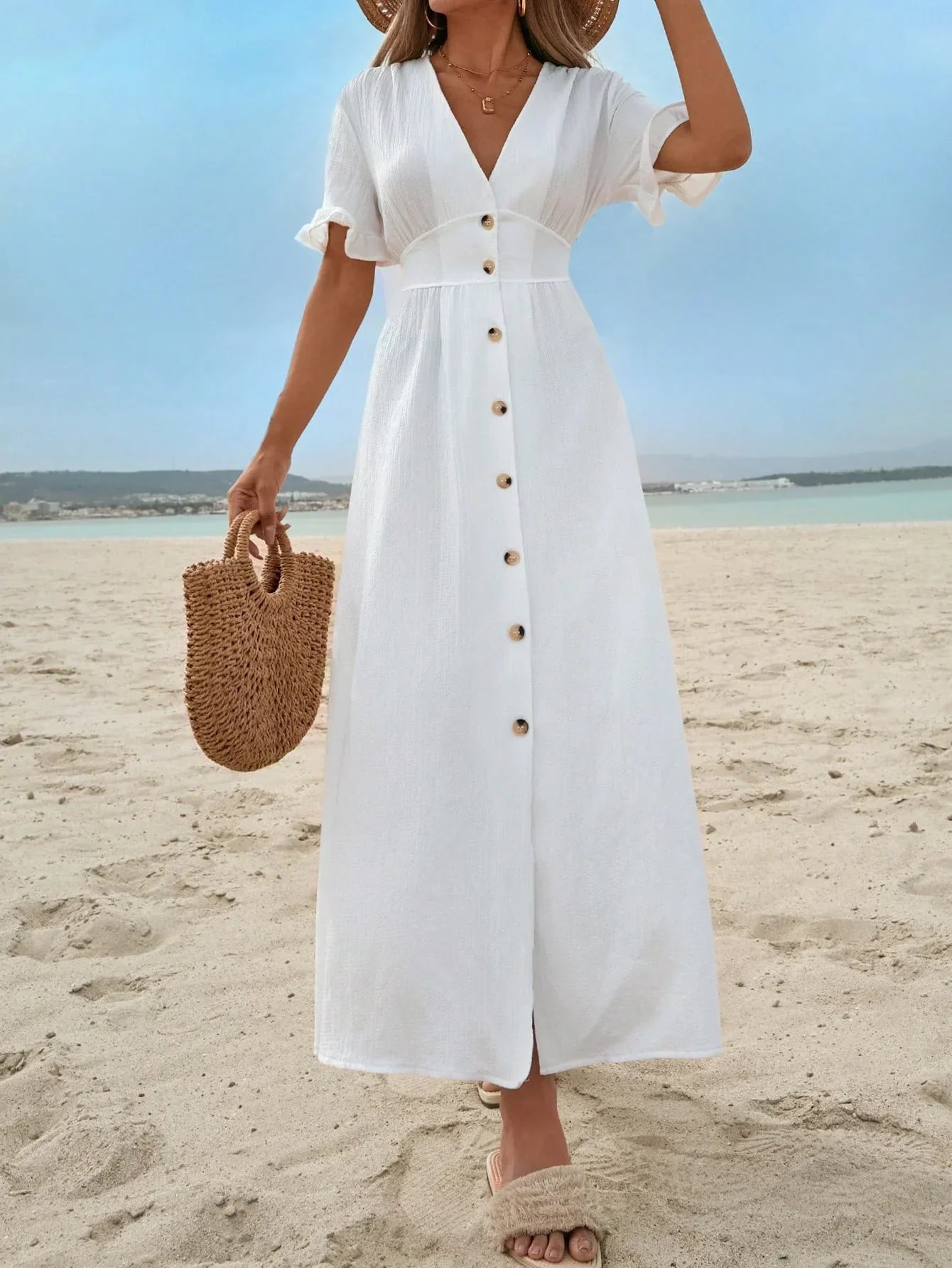 White - A woman on a sandy beach wearing a white textured crepe maxi dress with a V-neckline, flutter sleeves, button-down front, and a high leg slit, holding a woven straw handbag.
