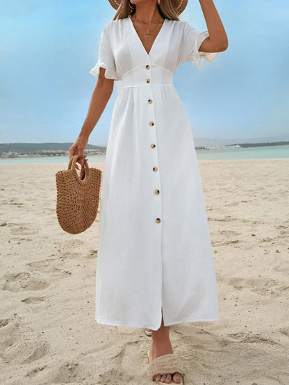 White - A woman on a sandy beach wearing a white textured crepe maxi dress with a V-neckline, flutter sleeves, button-down front, and a high leg slit, holding a woven straw handbag.