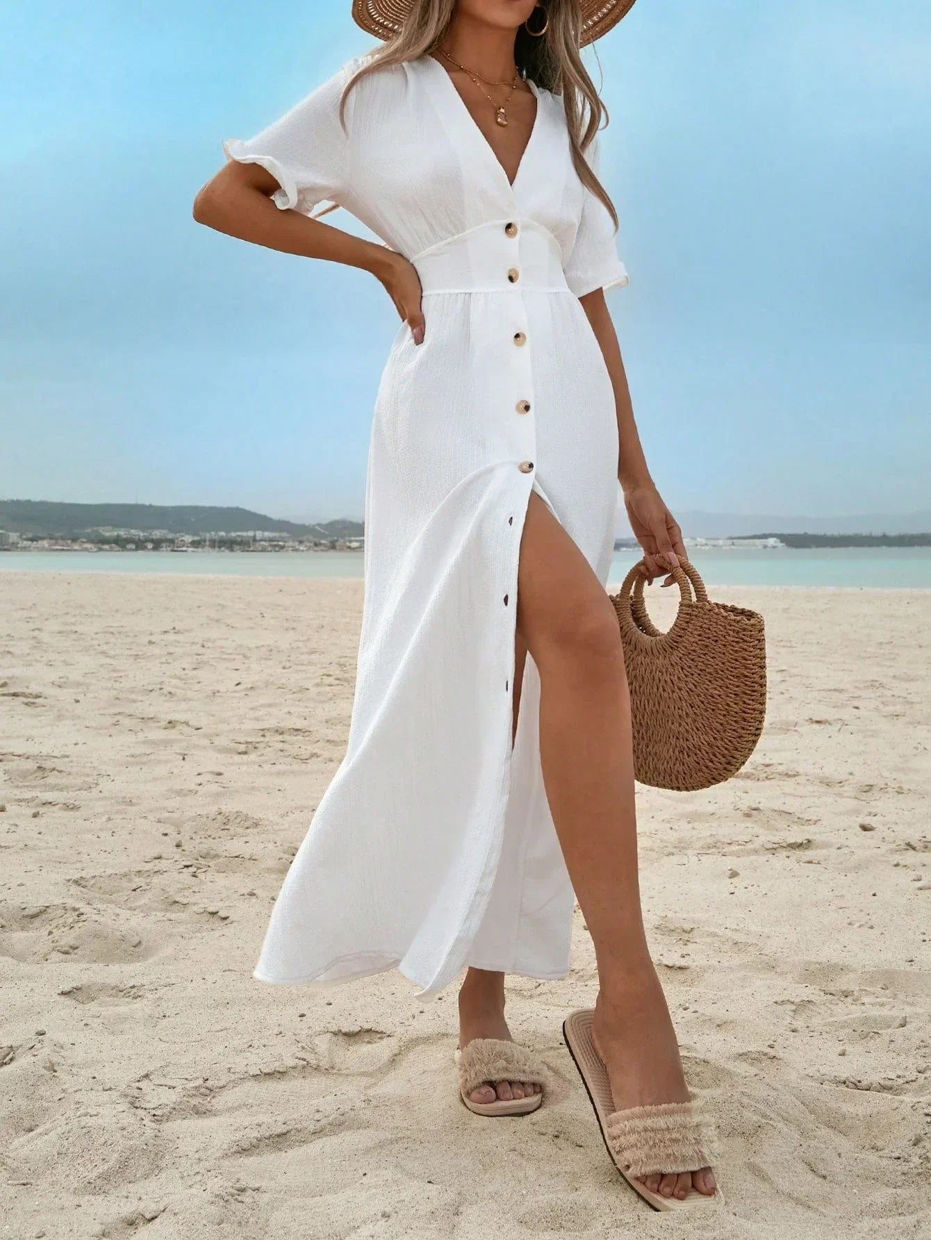 White - A woman on a sandy beach wearing a white textured crepe maxi dress with a V-neckline, flutter sleeves, button-down front, and a high leg slit, holding a woven straw handbag.