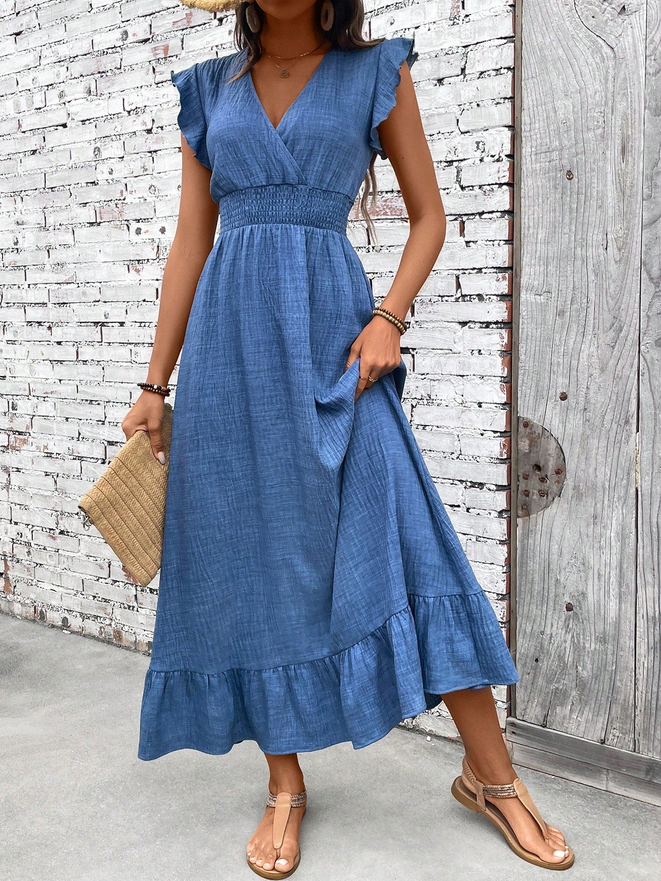Blue - A woman wearing a slate blue, slub-textured midi dress with flutter sleeves, a surplice V-neckline, a smocked waist, and a ruffled hem, styled with flat sandals and a straw clutch.