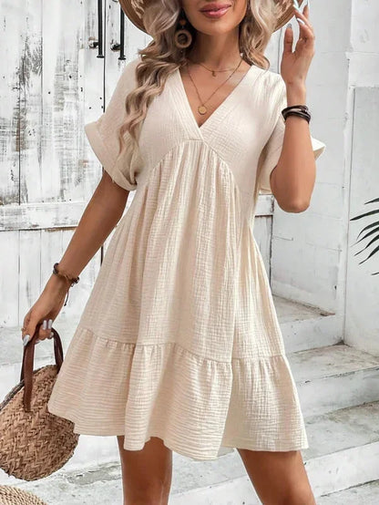 Light beige textured linen mini dress featuring a V-neckline, cuffed short sleeves, and a tiered ruffled hem.