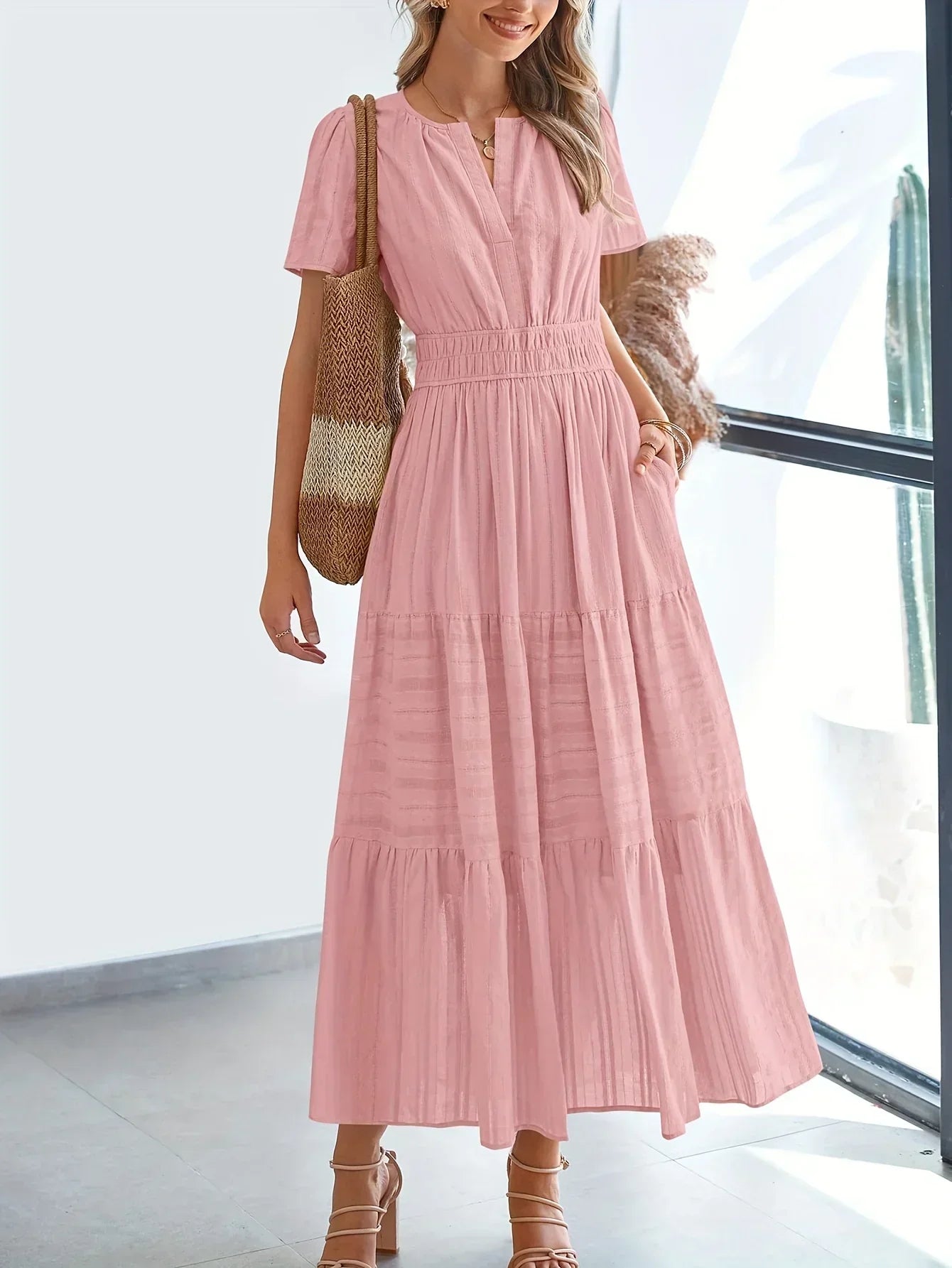 Pink - A woman wearing a dusty pink, textured stripe maxi dress featuring a smocked waist, tiered skirt, short sleeves, and a split neckline, styled with a woven straw shoulder bag.