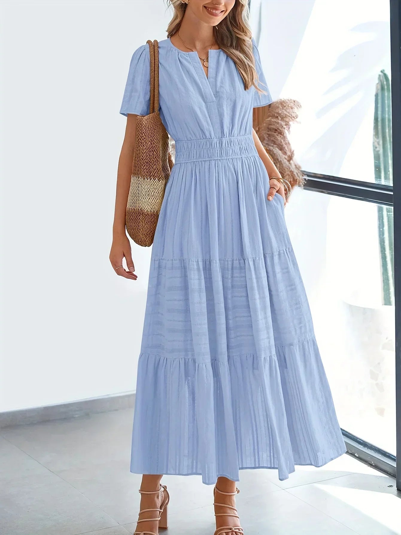 Light Blue - A woman wearing a dusty pink, textured stripe maxi dress featuring a smocked waist, tiered skirt, short sleeves, and a split neckline, styled with a woven straw shoulder bag.