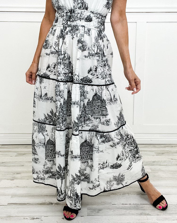 Black - A woman wearing a white tiered maxi dress with a brown toile de Jouy print, featuring short puff sleeves, a smocked waist, and a split neckline.