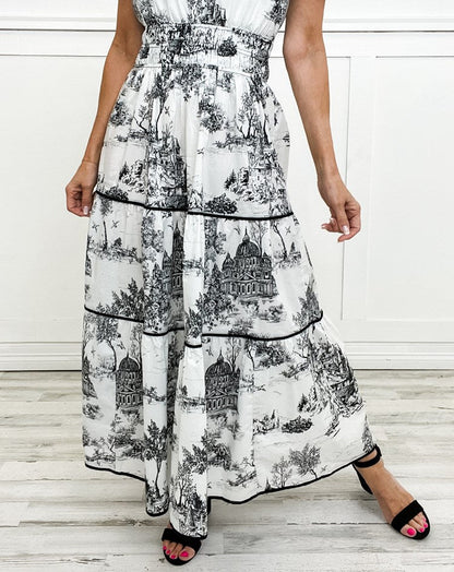 Black - A woman wearing a white tiered maxi dress with a brown toile de Jouy print, featuring short puff sleeves, a smocked waist, and a split neckline.