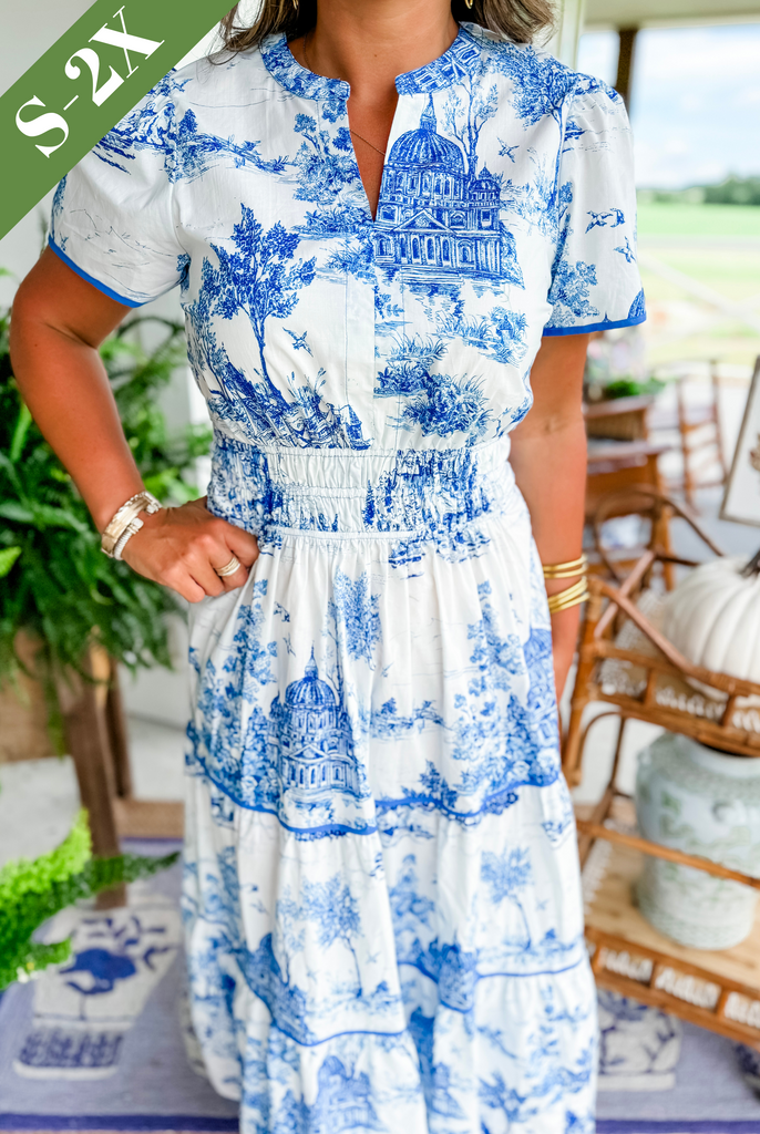Blue - A woman wearing a white tiered maxi dress with a brown toile de Jouy print, featuring short puff sleeves, a smocked waist, and a split neckline.