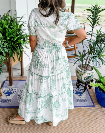 Blue - A woman wearing a white tiered maxi dress with a brown toile de Jouy print, featuring short puff sleeves, a smocked waist, and a split neckline.