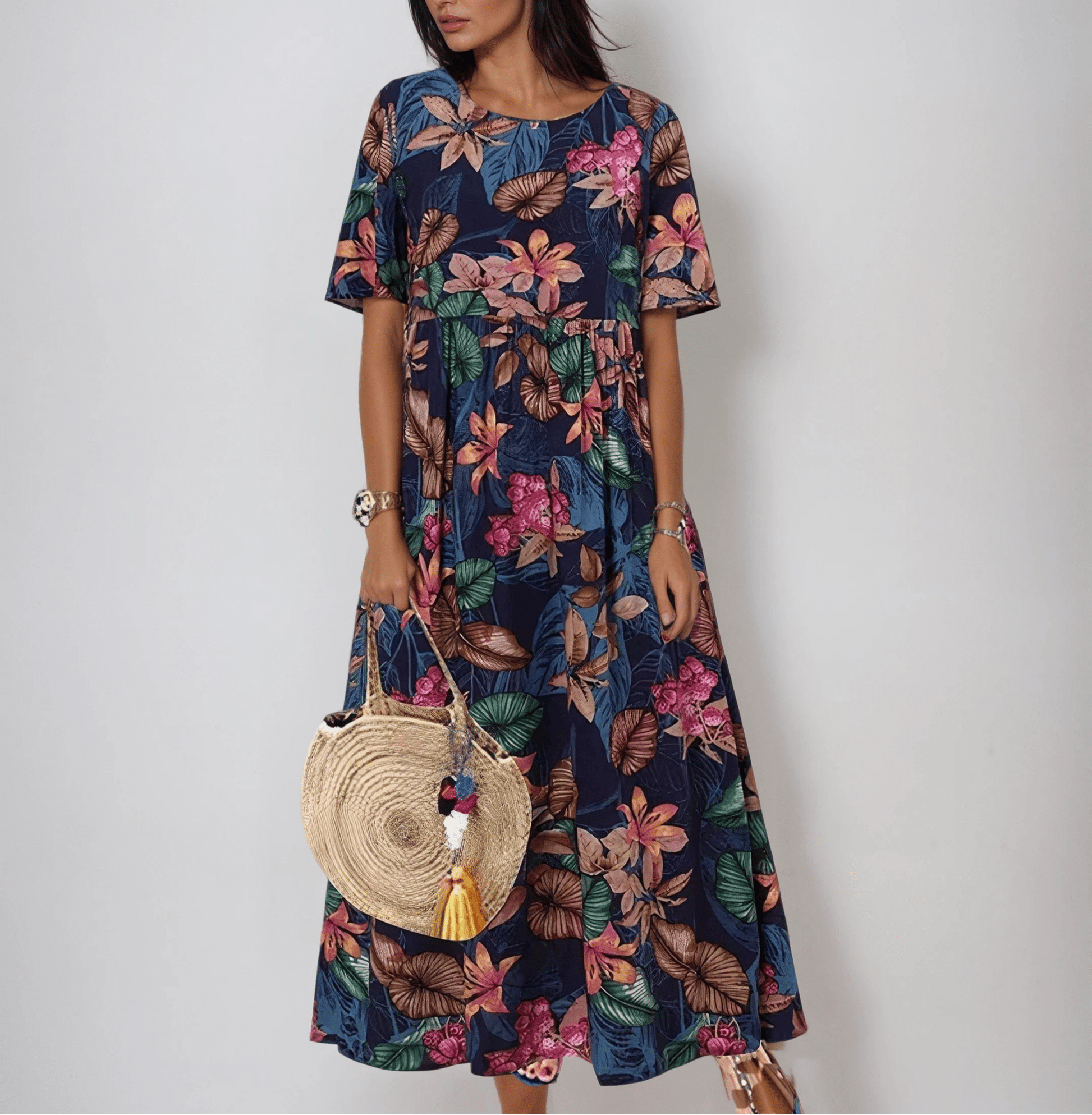 Blue - A woman wearing a navy blue maxi dress with a vibrant pink, orange, and green tropical floral print, featuring short sleeves, a crewneck, and an empire waist, holding a round woven straw bag.