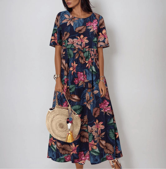 Blue - A woman wearing a navy blue maxi dress with a vibrant pink, orange, and green tropical floral print, featuring short sleeves, a crewneck, and an empire waist, holding a round woven straw bag.