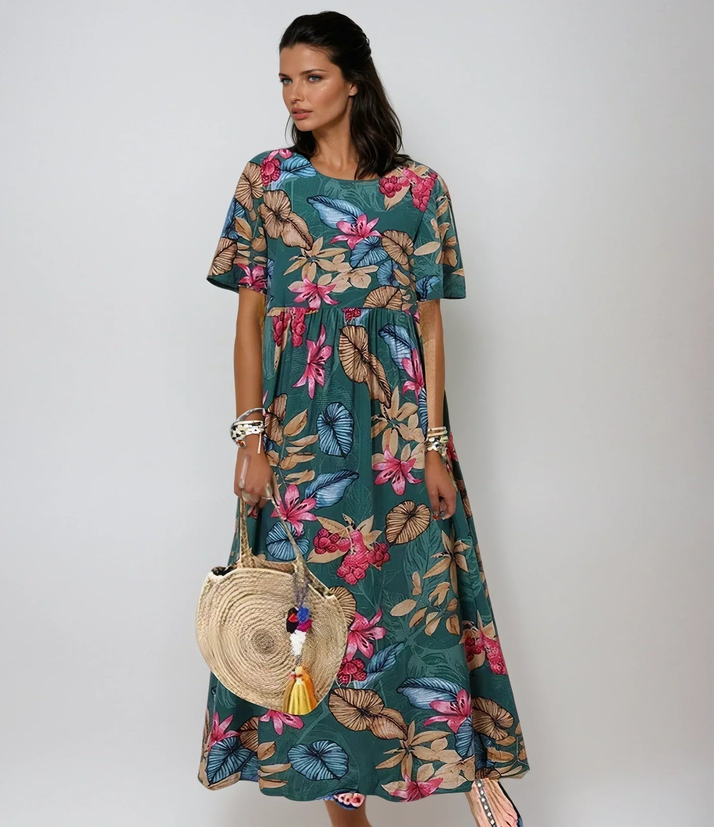 Green - A woman wearing a navy blue maxi dress with a vibrant pink, orange, and green tropical floral print, featuring short sleeves, a crewneck, and an empire waist, holding a round woven straw bag.