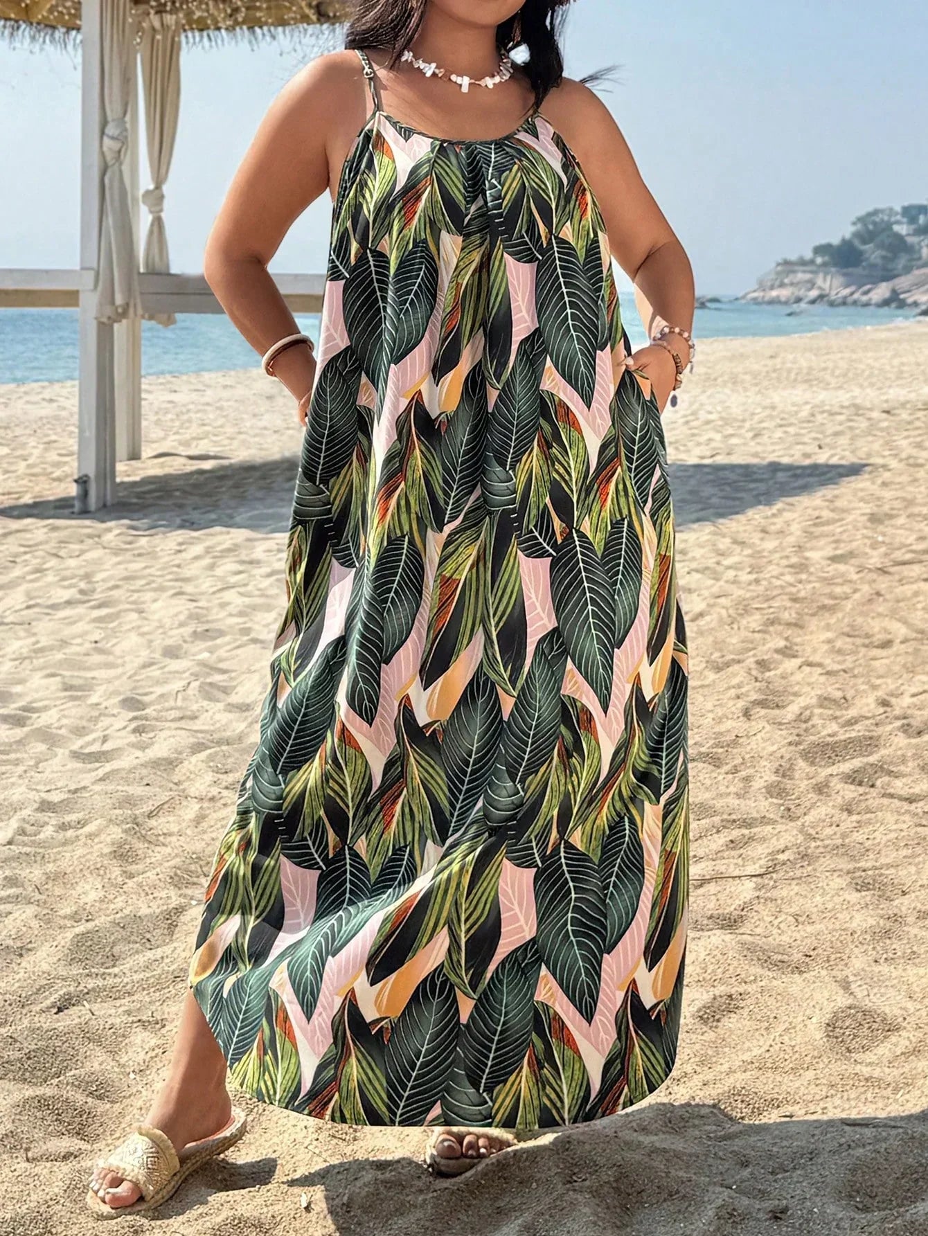 Versatile Colors - A woman stands on a sandy beach wearing a sleeveless maxi dress with a bold green, pink, and yellow tropical leaf print, featuring spaghetti straps and side pockets.