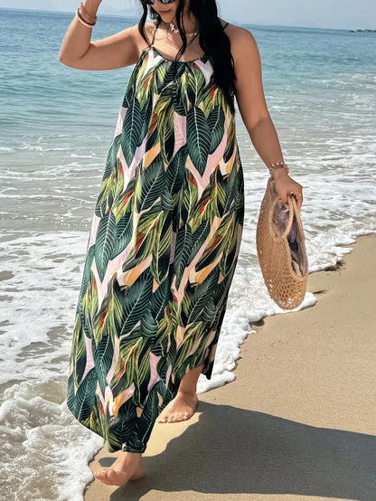 Versatile Colors - A woman stands on a sandy beach wearing a sleeveless maxi dress with a bold green, pink, and yellow tropical leaf print, featuring spaghetti straps and side pockets.