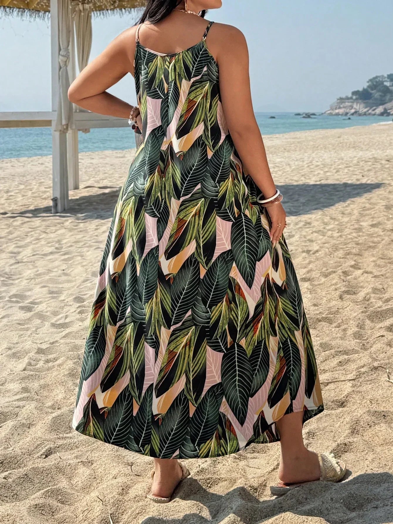 Versatile Colors - A woman stands on a sandy beach wearing a sleeveless maxi dress with a bold green, pink, and yellow tropical leaf print, featuring spaghetti straps and side pockets.