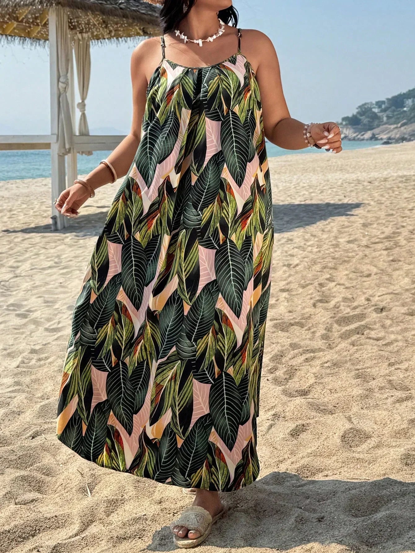 Versatile Colors - A woman stands on a sandy beach wearing a sleeveless maxi dress with a bold green, pink, and yellow tropical leaf print, featuring spaghetti straps and side pockets.