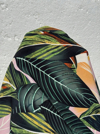 Versatile Colors - A woman stands on a sandy beach wearing a sleeveless maxi dress with a bold green, pink, and yellow tropical leaf print, featuring spaghetti straps and side pockets.