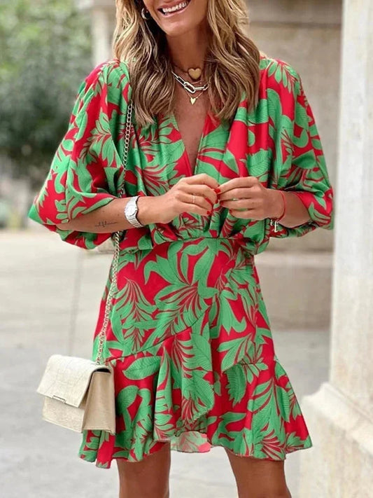 Woman wearing a red mini dress with a green tropical leaf print, featuring a deep V-neckline, dolman sleeves, and an asymmetrical ruffle hem.