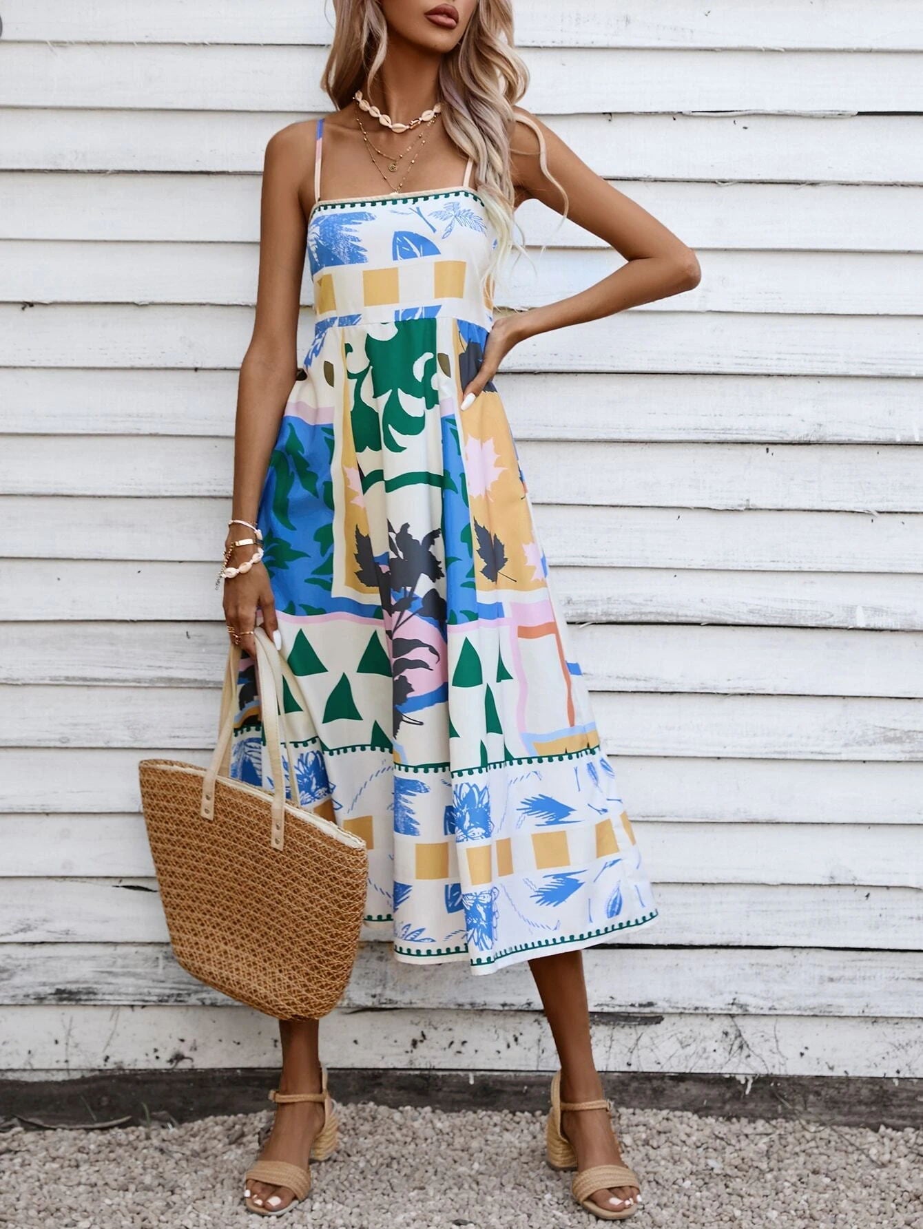 Beige - A woman wearing a white midi dress with a colorful tropical and geometric patchwork print, featuring a square neckline and thin straps, paired with a woven straw tote bag and neutral block heel sandals against a white wooden wall.