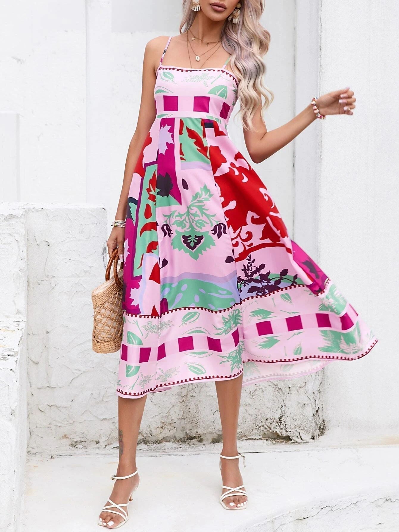 Rose Red - A woman wearing a white midi dress with a colorful tropical and geometric patchwork print, featuring a square neckline and thin straps, paired with a woven straw tote bag and neutral block heel sandals against a white wooden wall.