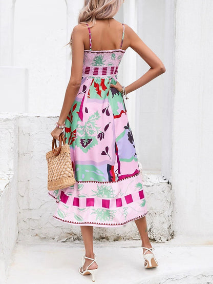 Rose Red - A woman wearing a white midi dress with a colorful tropical and geometric patchwork print, featuring a square neckline and thin straps, paired with a woven straw tote bag and neutral block heel sandals against a white wooden wall.