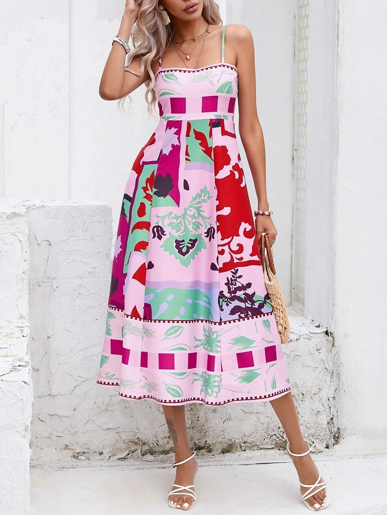 Rose Red - A woman wearing a white midi dress with a colorful tropical and geometric patchwork print, featuring a square neckline and thin straps, paired with a woven straw tote bag and neutral block heel sandals against a white wooden wall.