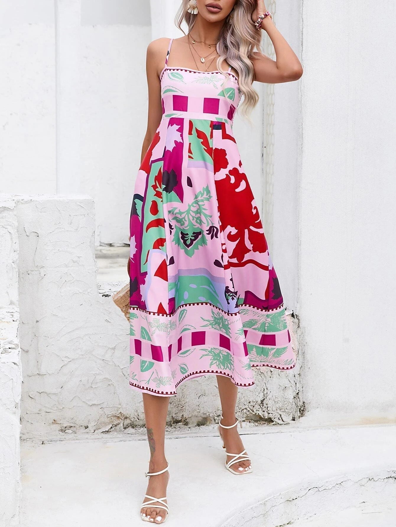 Rose Red - A woman wearing a white midi dress with a colorful tropical and geometric patchwork print, featuring a square neckline and thin straps, paired with a woven straw tote bag and neutral block heel sandals against a white wooden wall.