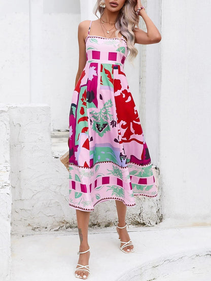 Rose Red - A woman wearing a white midi dress with a colorful tropical and geometric patchwork print, featuring a square neckline and thin straps, paired with a woven straw tote bag and neutral block heel sandals against a white wooden wall.