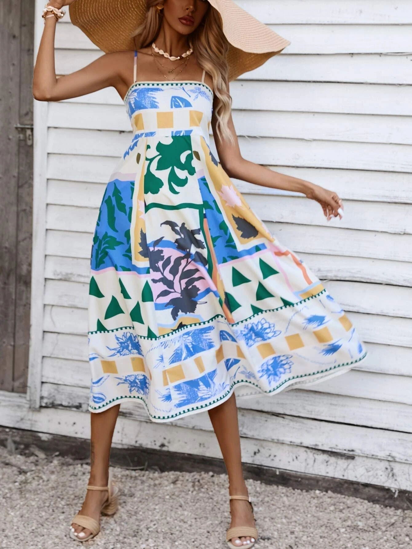 Beige - A woman wearing a white midi dress with a colorful tropical and geometric patchwork print, featuring a square neckline and thin straps, paired with a woven straw tote bag and neutral block heel sandals against a white wooden wall.