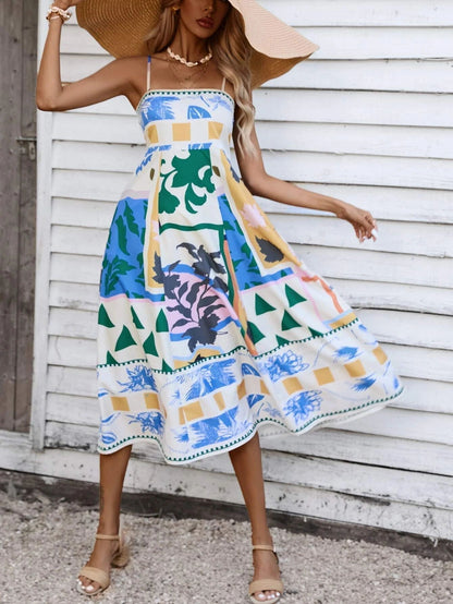 Beige - A woman wearing a white midi dress with a colorful tropical and geometric patchwork print, featuring a square neckline and thin straps, paired with a woven straw tote bag and neutral block heel sandals against a white wooden wall.
