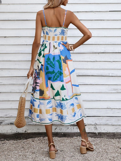 Beige - A woman wearing a white midi dress with a colorful tropical and geometric patchwork print, featuring a square neckline and thin straps, paired with a woven straw tote bag and neutral block heel sandals against a white wooden wall.