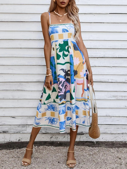 Beige - A woman wearing a white midi dress with a colorful tropical and geometric patchwork print, featuring a square neckline and thin straps, paired with a woven straw tote bag and neutral block heel sandals against a white wooden wall.