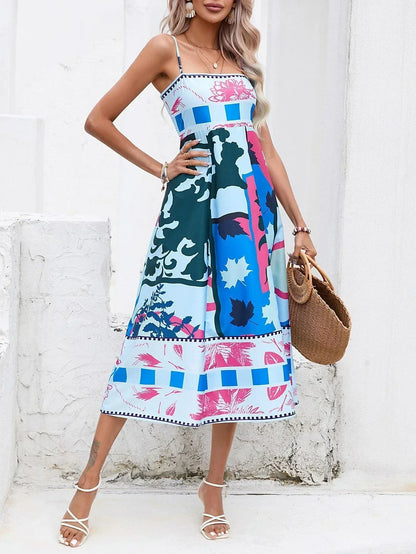 Blue - A woman wearing a white midi dress with a colorful tropical and geometric patchwork print, featuring a square neckline and thin straps, paired with a woven straw tote bag and neutral block heel sandals against a white wooden wall.