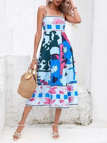Blue - A woman wearing a white midi dress with a colorful tropical and geometric patchwork print, featuring a square neckline and thin straps, paired with a woven straw tote bag and neutral block heel sandals against a white wooden wall.